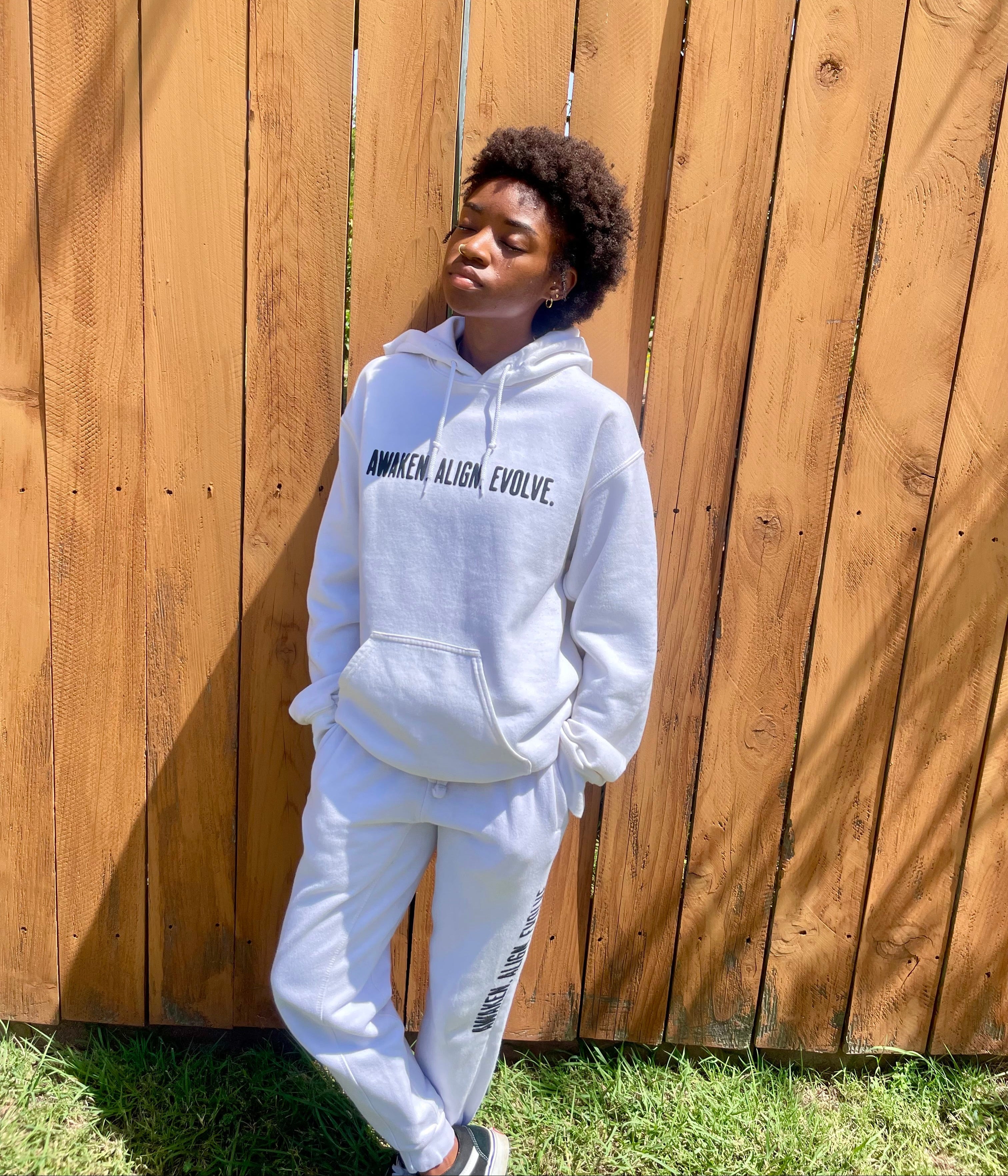 A person standing outdoors in front of a wooden fence wearing a white hoodie with black text reading 'AWAKEN ALIGN EVOLVE' and matching white sweatpants.