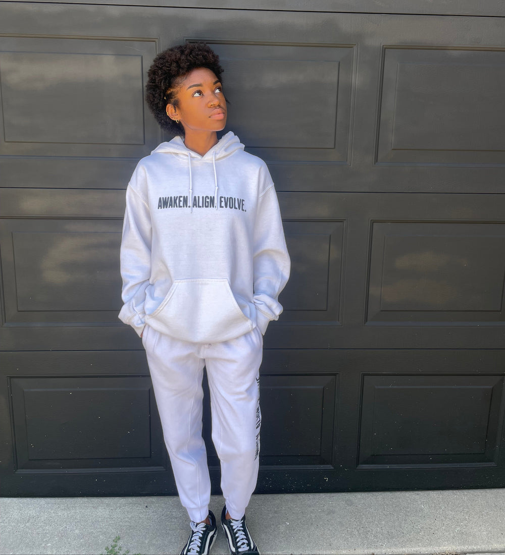 A person wearing a white hoodie and matching sweatpants stands in front of a black paneled garage door. The hoodie has black text on the chest that reads 'AWAKEN. ALIGN. EVOLVE'.