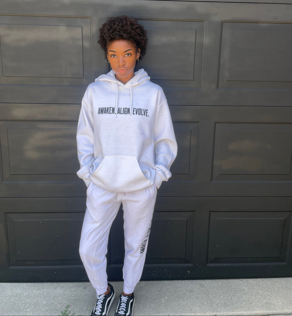 A person wearing a white hoodie and matching sweatpants stands in front of a black paneled garage door. The hoodie has black text on the chest that reads 'AWAKEN. ALIGN. EVOLVE'.