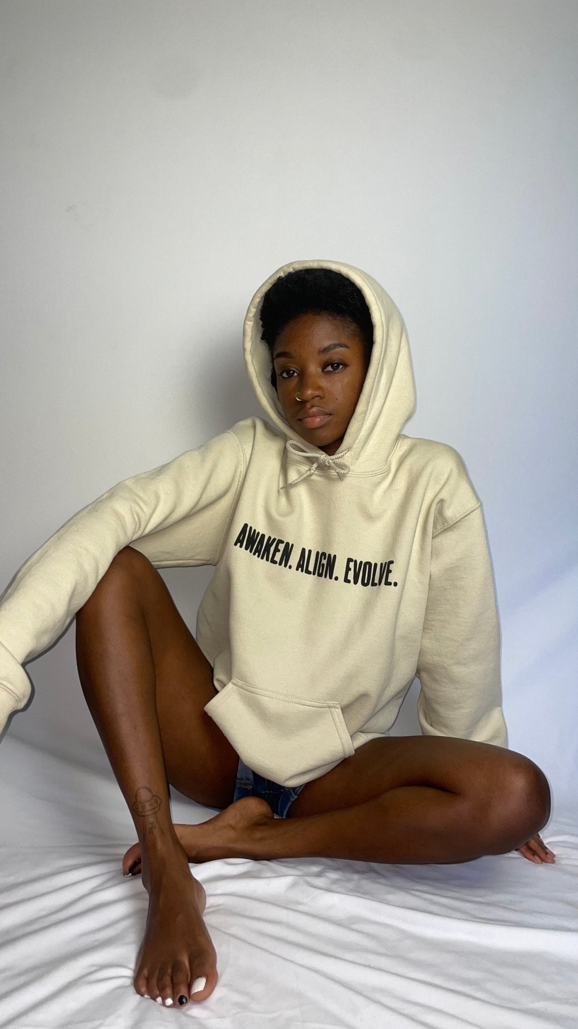 Person wearing a beige hoodie with text "AWAKEN.ALIGN.EVOLVE.' sitting on a white surface.
